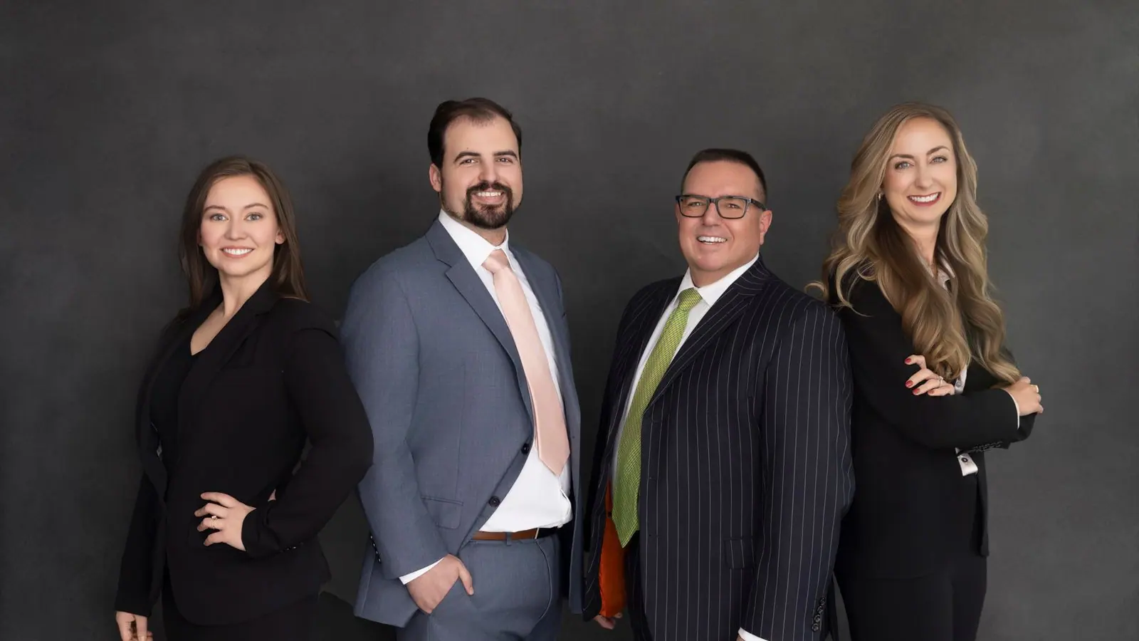 Four people in different tailored outfits photographed against a dark gray studio backdrop